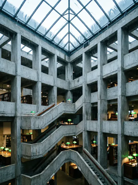 A symmetrical brutalist concrete library atrium with tiered balconies, green desk lamps and a glass roof streaked with rain.