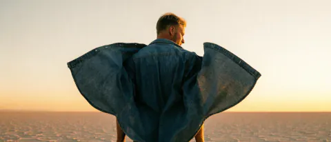 A man seen from behind on a salt flat at sunrise, his denim shirt blown out wide on both sides like wings.