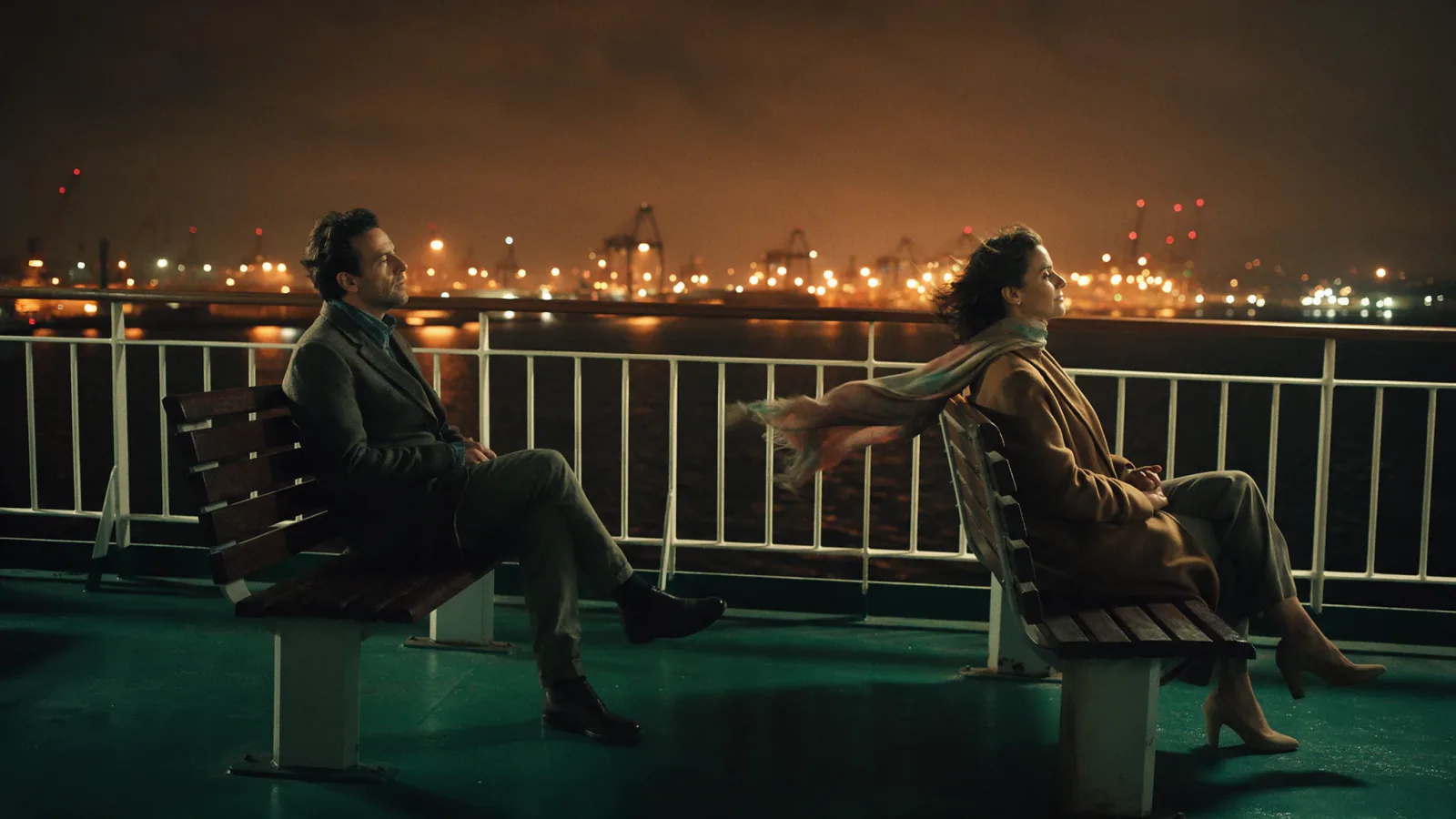 A man and a woman sit on separate benches on a night ferry deck, her scarf blowing toward him against orange harbour lights.