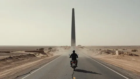 A lone motorcyclist rides a cracked desert highway toward a towering black monolith in pale morning haze.